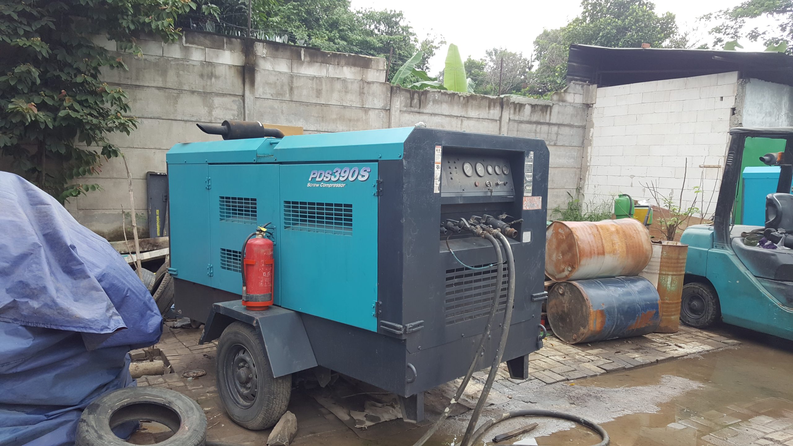 Unit kompresor diesel PDS390S berkapasitas tinggi siap digunakan di area proyek, lengkap dengan fire extinguisher dan selang tekanan tinggi. Terlihat lingkungan kerja khas lapangan industri dengan drum oli, forklift, dan suasana outdoor yang menunjukkan kesiapan operasional.