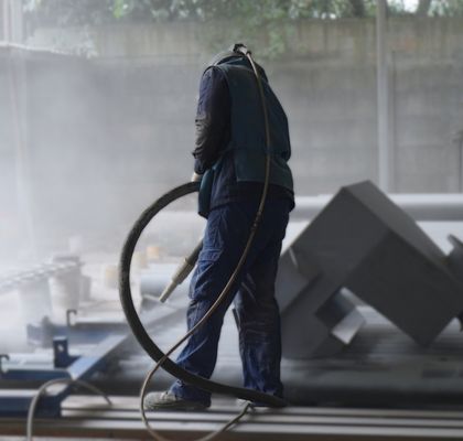 Seorang pekerja sandblasting mengenakan alat pelindung diri (APD) lengkap, termasuk helm khusus, sedang membersihkan permukaan tangki metal besar di lingkungan industri.